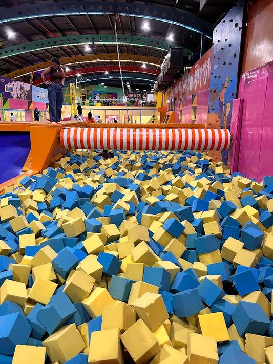 A young woman stands on the edge of a foam pit filled with blue and yellow cubes at an indoor trampoline park.