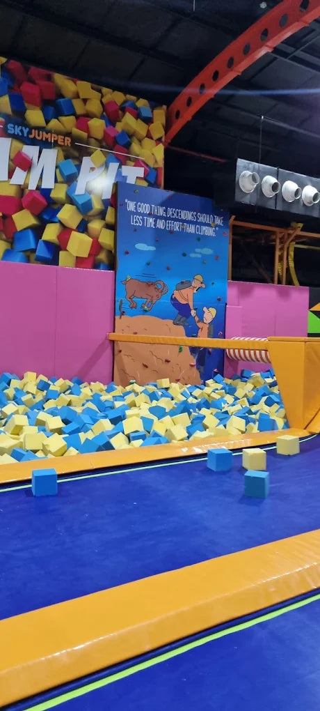 A foam pit filled with blue and yellow cubes at a trampoline park, with a climbing wall and a motivational quote.