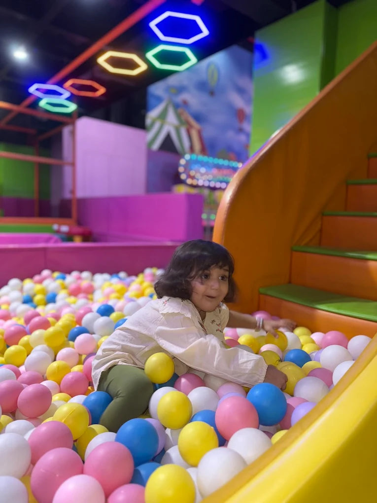 A young child plays in a ball pit with colorful balls, with a slide and neon lights in the background.