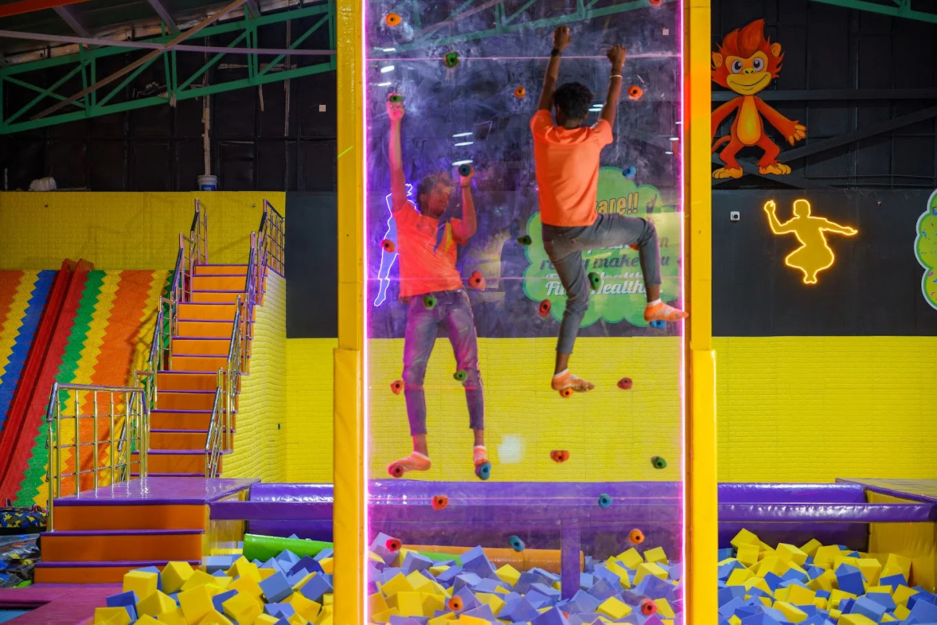 Two young men climb a colorful indoor rock wall at a trampoline park.