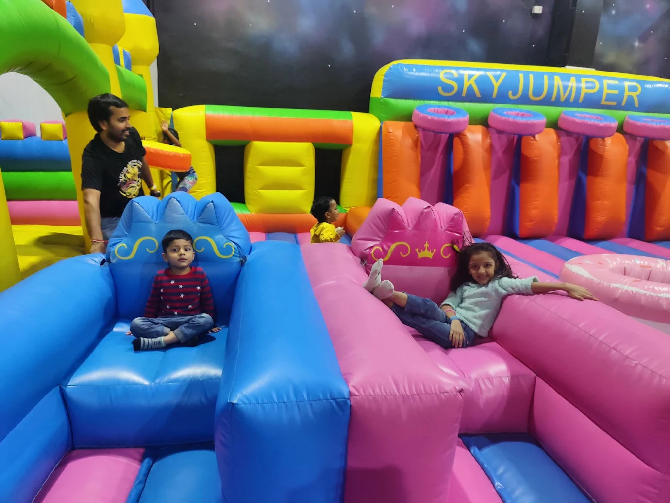Children play on a colorful inflatable obstacle course at an indoor playground. A man supervises from the side.