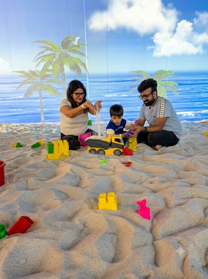 A family plays in a sandpit with beach-themed projections. A child is focused on a toy truck while parents supervise.