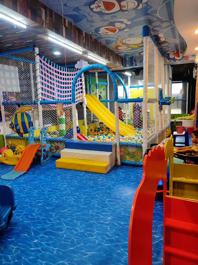 Children play in a colorful indoor playground with ball pits and slides. The ceiling is decorated with cartoon characters.
