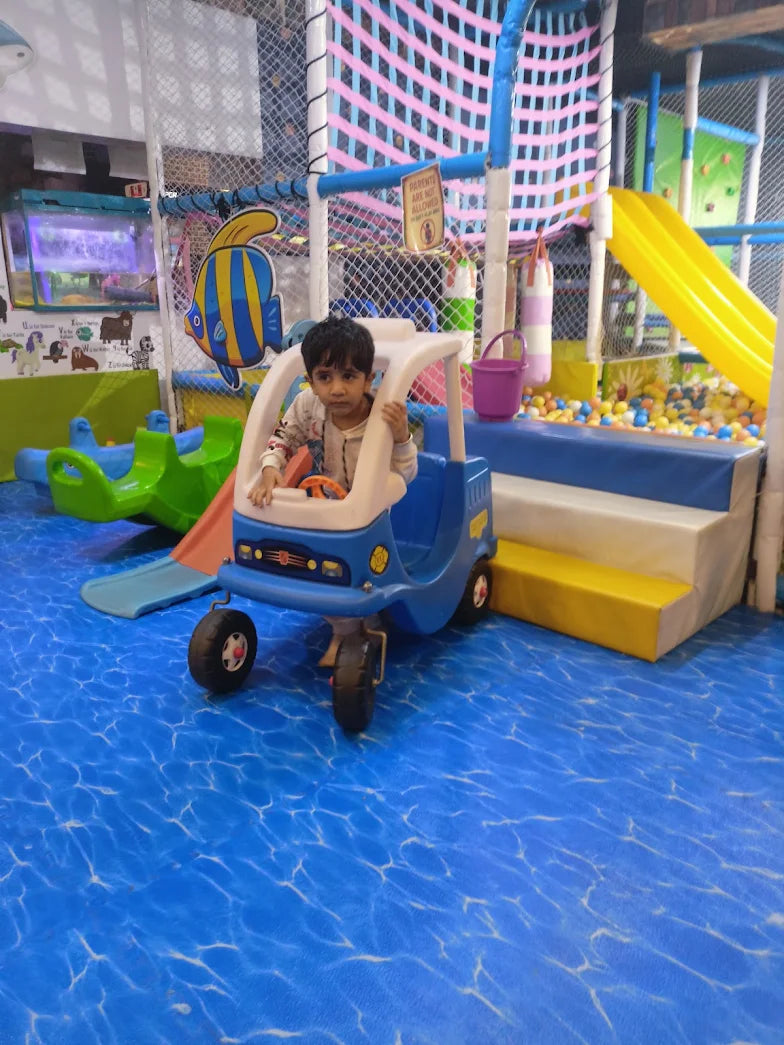 A young child sits in a toy car in a colorful indoor playground with a ball pit and slide.