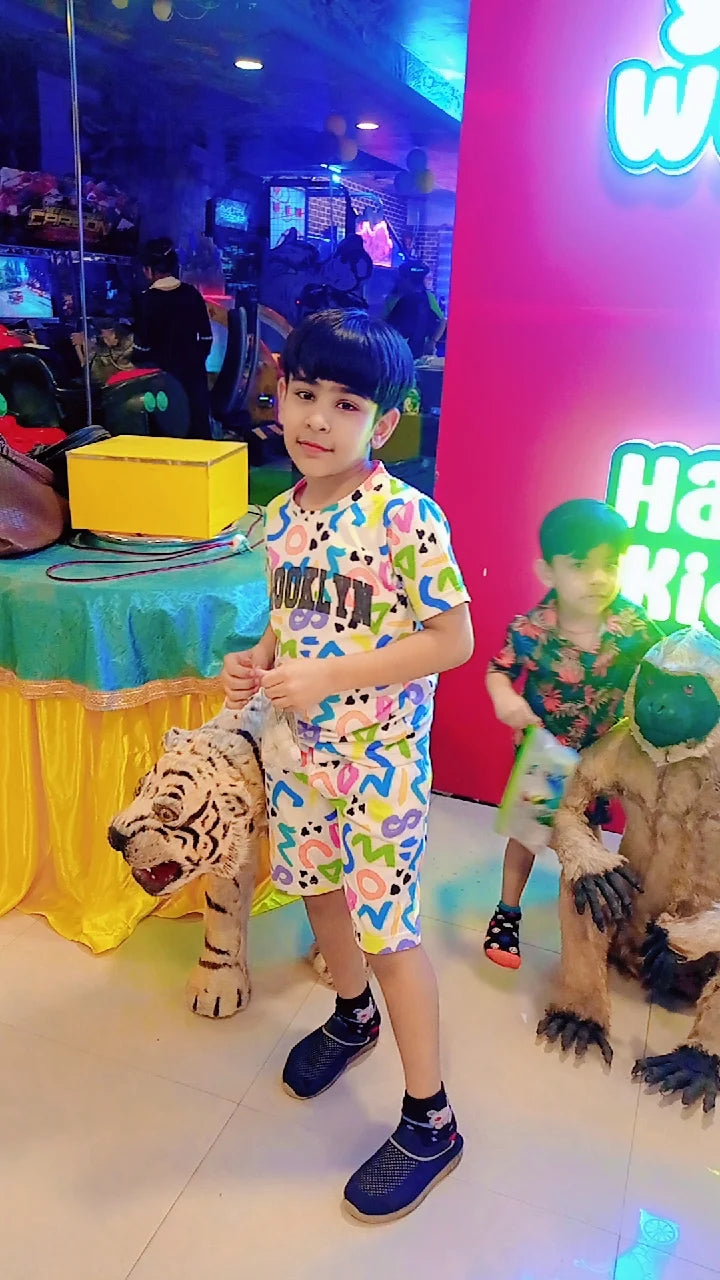 A young boy in colorful pajamas stands next to a tiger toy in an arcade. Another boy is visible in the background.