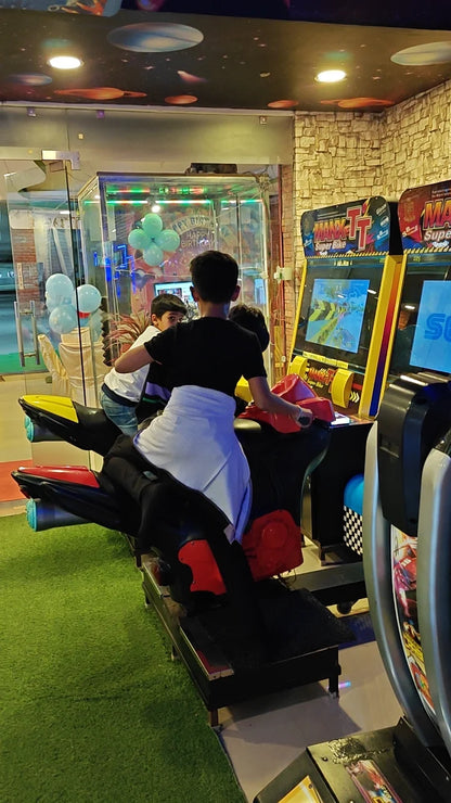 Children play a motorcycle racing arcade game in an entertainment center. The ceiling is decorated with planets and stars.
