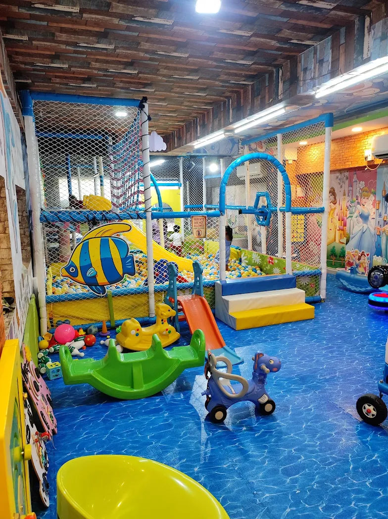 A colorful indoor playground with ball pits, slides, and ride-on toys for young children. The floor is blue with a water pattern, and the walls are decorated with cartoon characters.