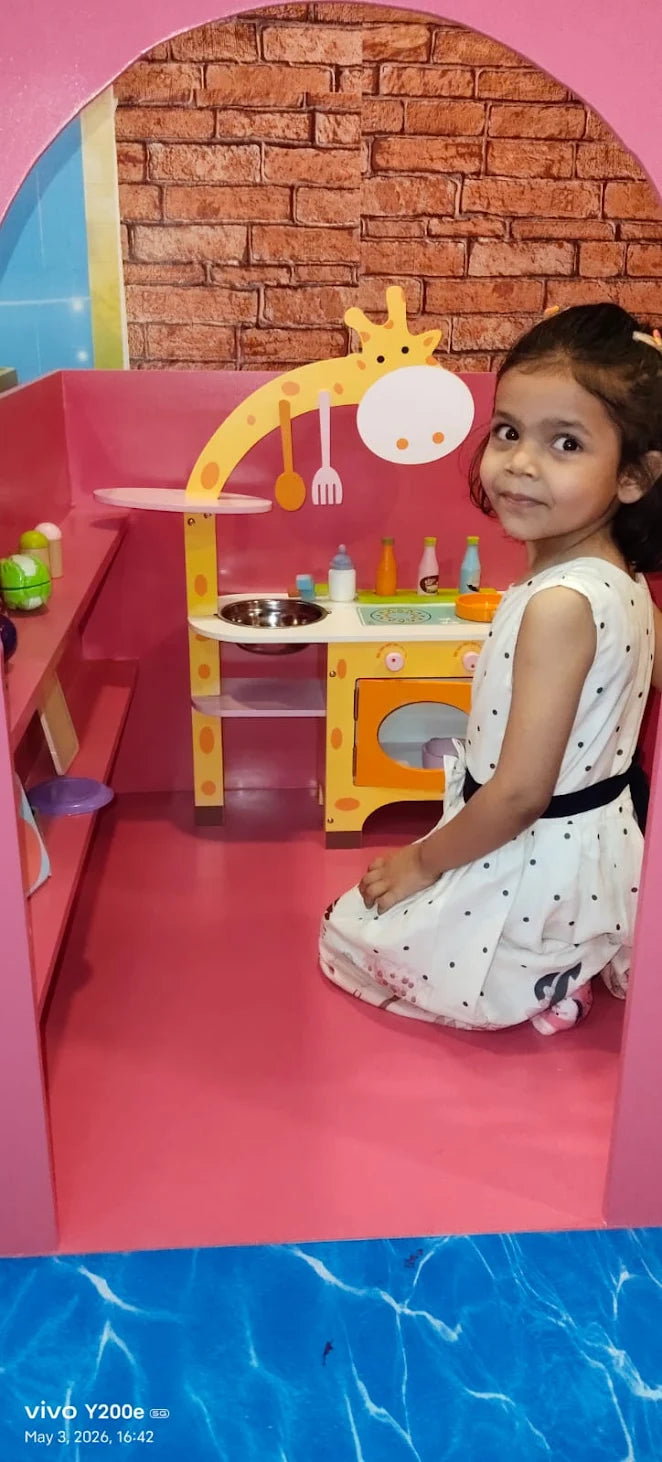A young girl plays in a toy kitchen with a giraffe theme. The kitchen is pink and yellow, with toy food and utensils.