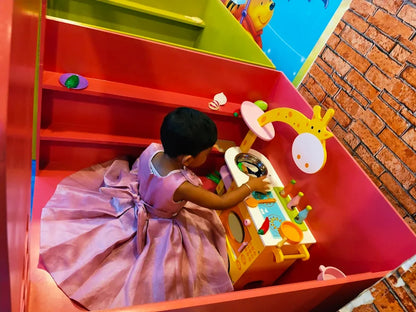 A young child in a pink dress plays with a toy kitchen in a colorful play area.