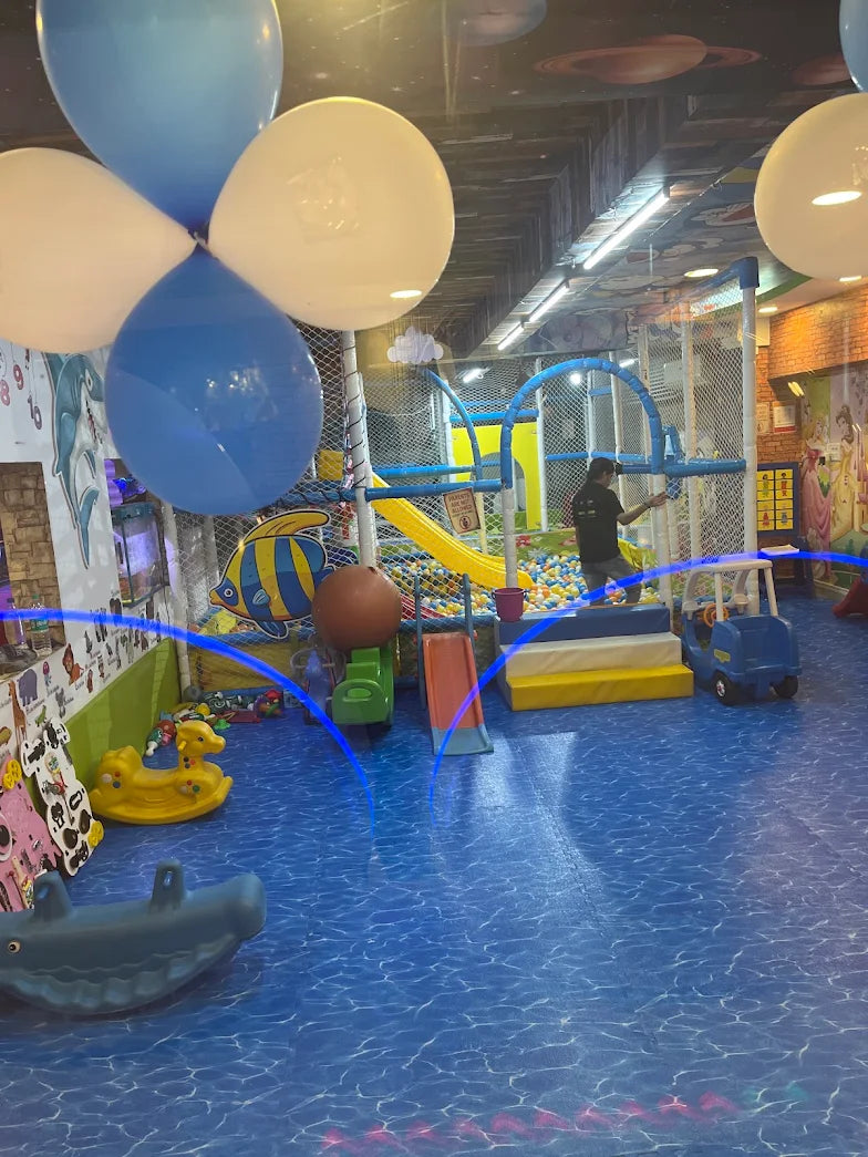 A colorful indoor playground with a ball pit, slides, and toys, decorated with balloons and cartoon characters.