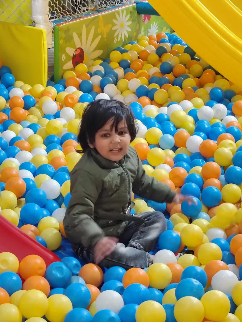 A young child sits in a ball pit filled with colorful balls, looking at the camera.