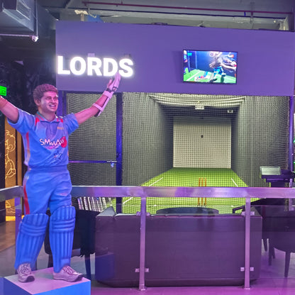 A statue of a cricketer stands in front of a batting cage with a screen showing a game.