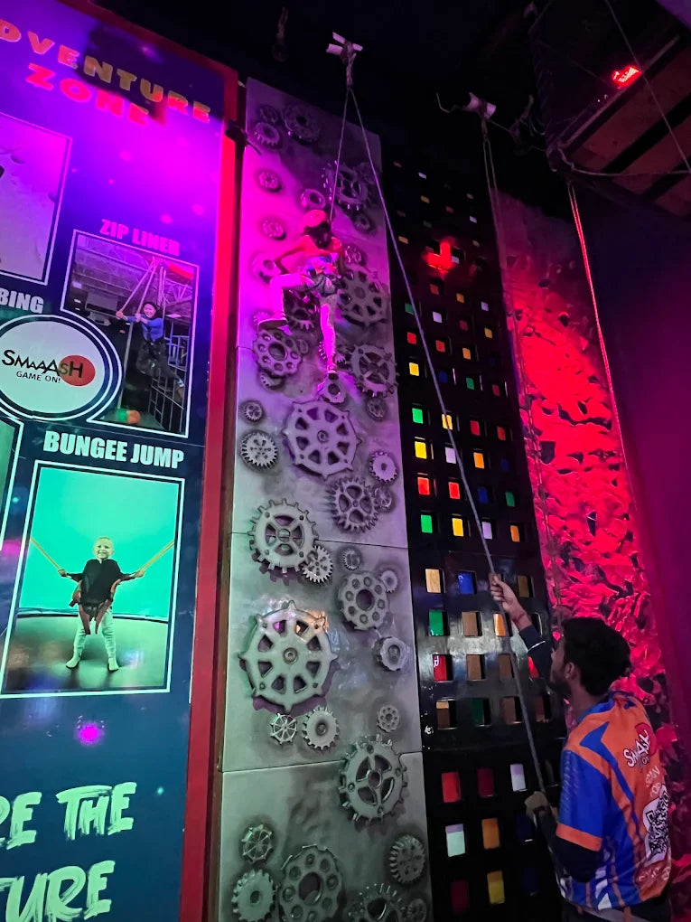 A young person climbs a gear-themed wall while a young adult belays them.