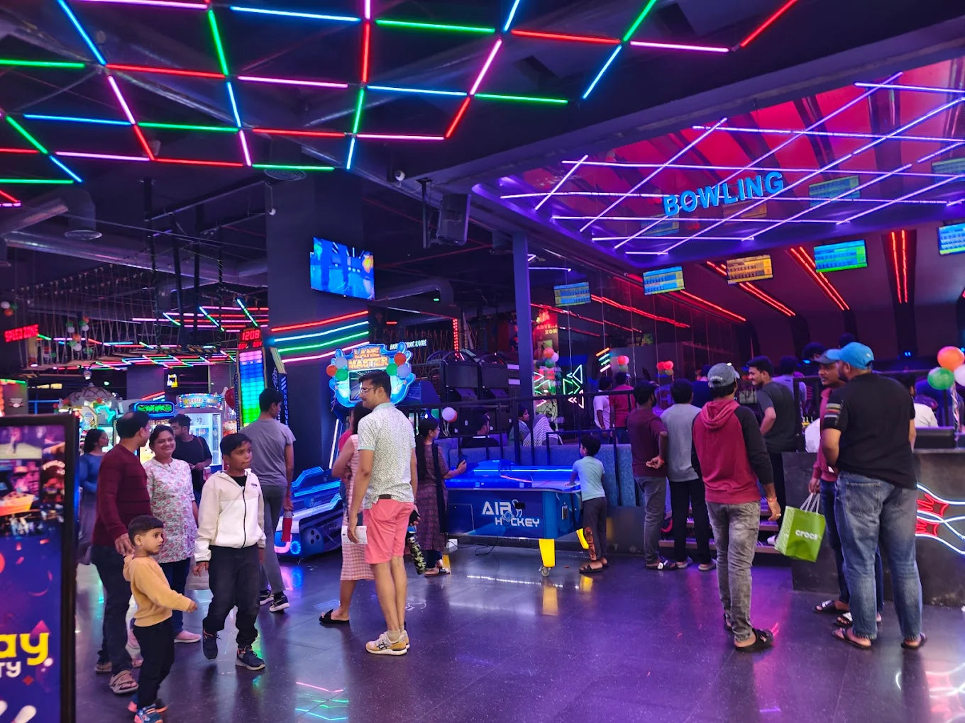 People of various ages enjoy an arcade with neon lights and games like bowling and air hockey.