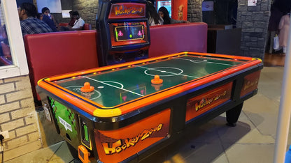 An air hockey table with a "Hockey 2000" design is in the foreground, with people in the background.