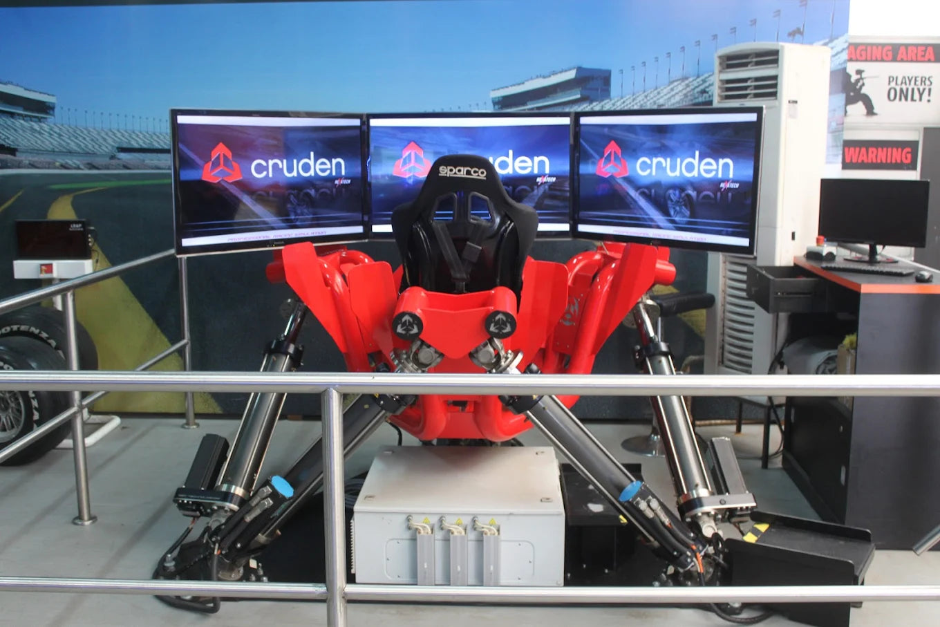A red racing simulator with three screens displays the "Cruden" logo, set up for players.