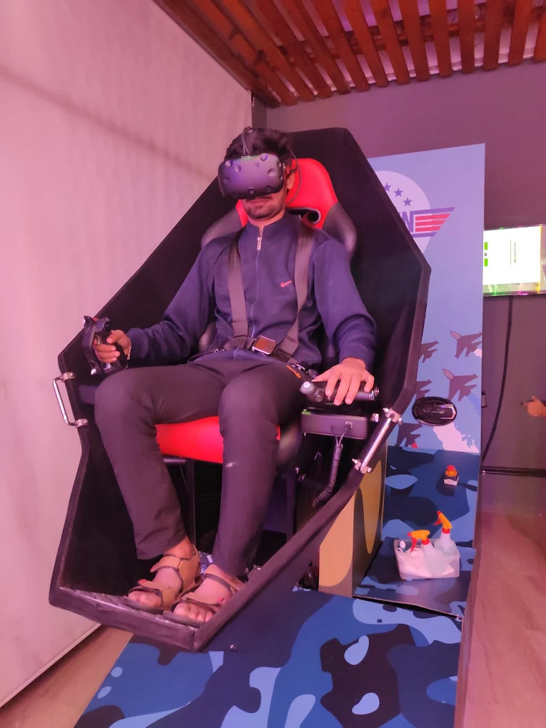 A young person wearing a VR headset sits in a flight simulator chair, holding controllers. The simulator has a camouflage design and is set up for an immersive experience.