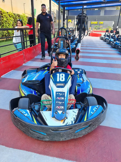 A young child in a go-kart gives a thumbs-up, with other go-karts and people in the background at an outdoor track.