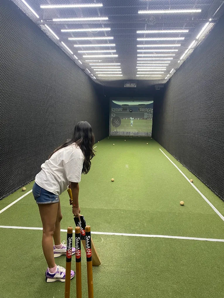 A young person practices cricket in an indoor batting cage with a projected screen.
