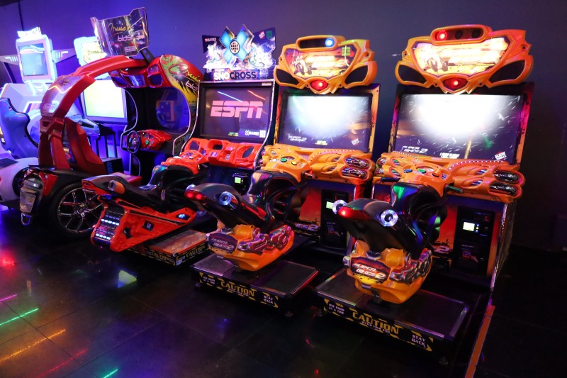 Arcade with multiple racing and shooting video games, some with motorcycle and car themes, illuminated by neon lights.