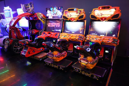Arcade with multiple racing and shooting video games, some with motorcycle and car themes, illuminated by neon lights.