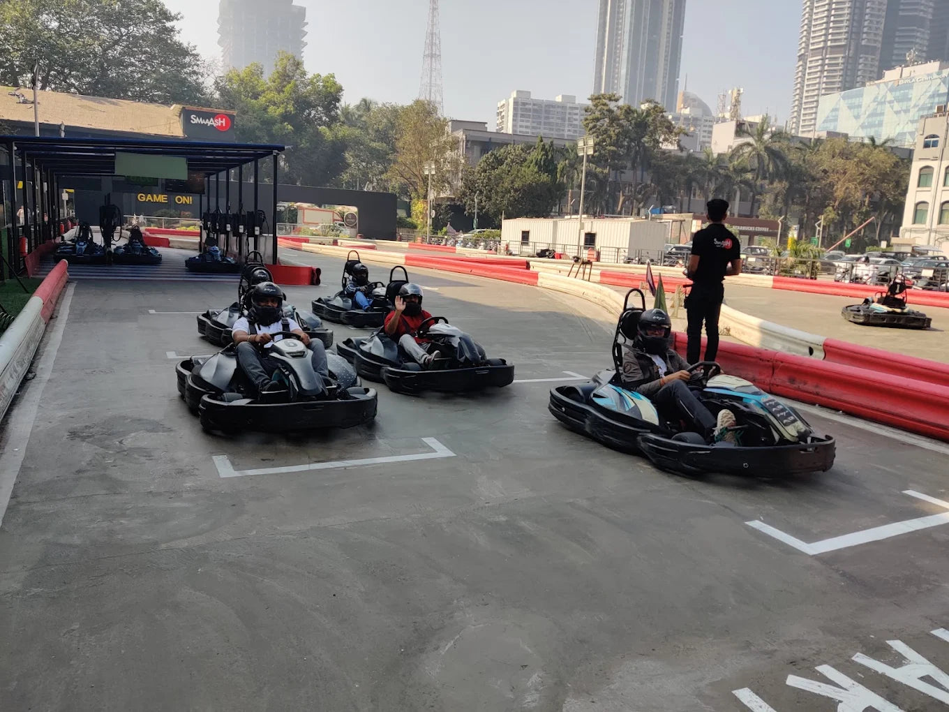 Adults race go-karts on an outdoor track with city buildings in the background.