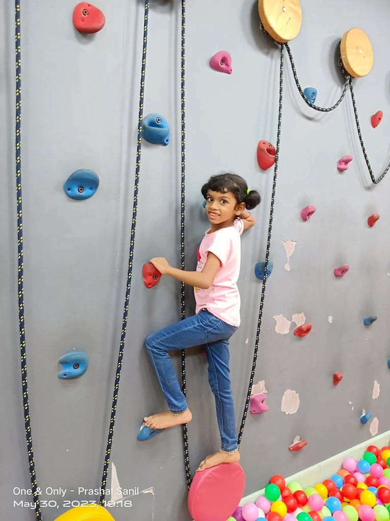 A young girl climbs a rock wall with colorful holds, while a ball pit is visible below.