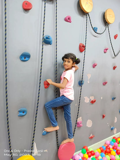 A young girl climbs a rock wall with colorful holds, while a ball pit is visible below.