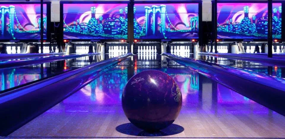 A purple bowling ball sits on a lane in a bowling alley with neon lights and digital screens.