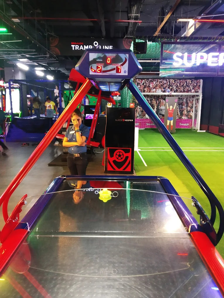 A young woman stands by an air hockey table in an arcade with a soccer-themed backdrop. The scoreboard shows the score as 2-7.