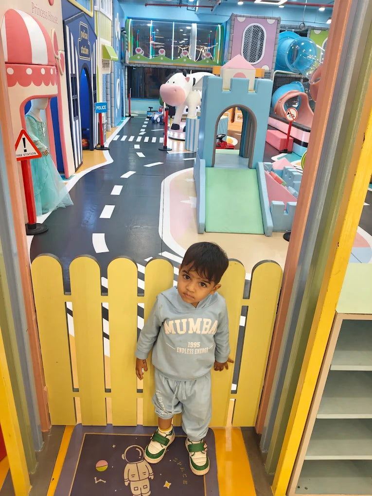 A young boy stands in a colorful indoor playground with a castle and play structures. He is wearing a light blue tracksuit.