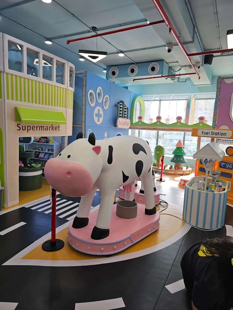 A child plays in a colorful indoor playground with a large cow statue and miniature shops.