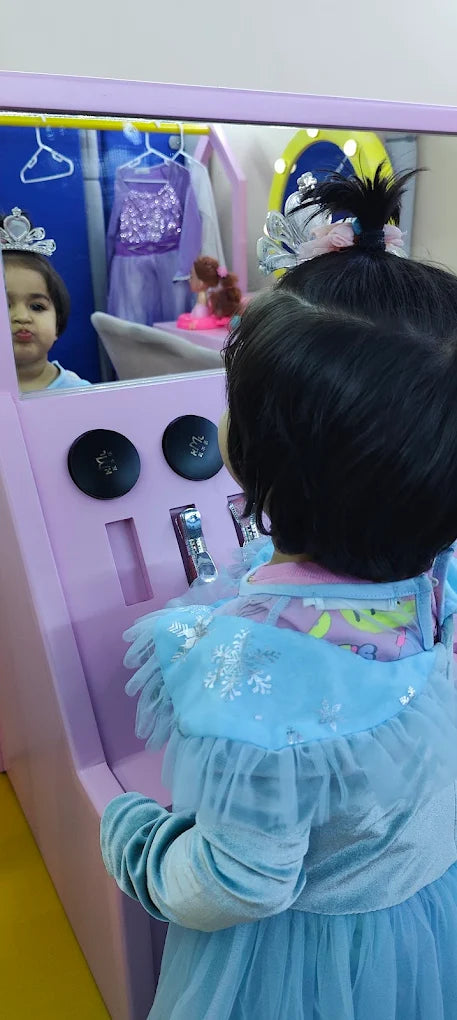 A young child in a blue dress looks in a mirror at a play vanity.