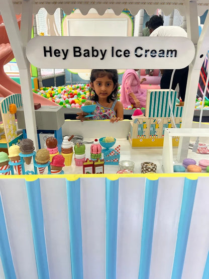A young girl stands behind an ice cream stand in a ball pit, holding a toy ice cream cone.