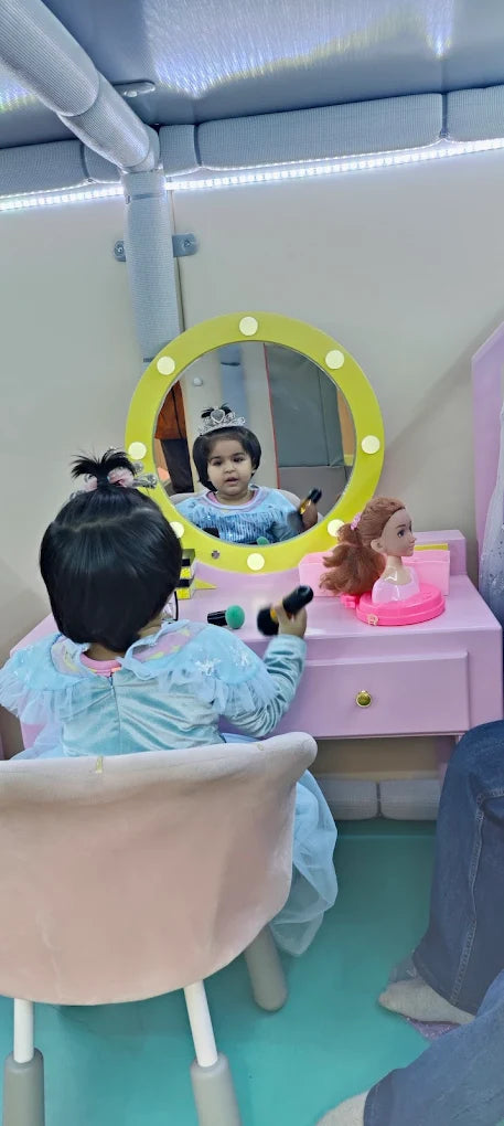 A young child in a princess dress plays with makeup at a vanity, reflected in a mirror.