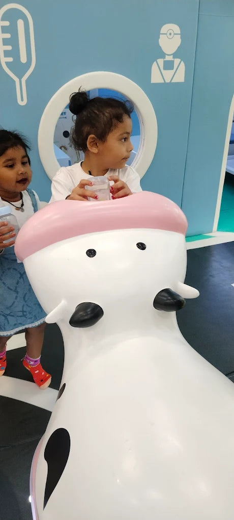 Two young children interact with a large, white cow-shaped toy in a brightly colored play area.