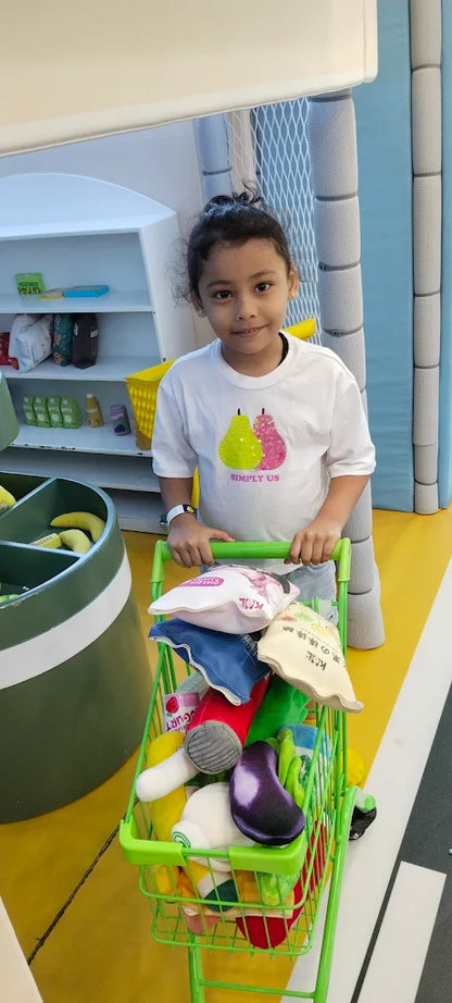 A young child pushes a toy shopping cart filled with plush food items in a play grocery store.