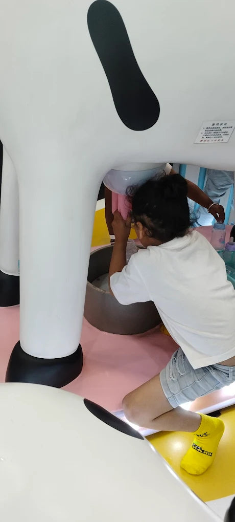A young girl kneels to "milk" a large, white cow-shaped toy in a colorful play area.