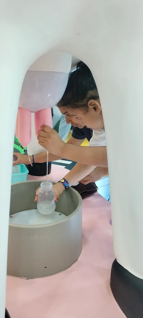 A person milks a large, pink udder-like structure into a bottle. The person is in a playful, interactive exhibit.