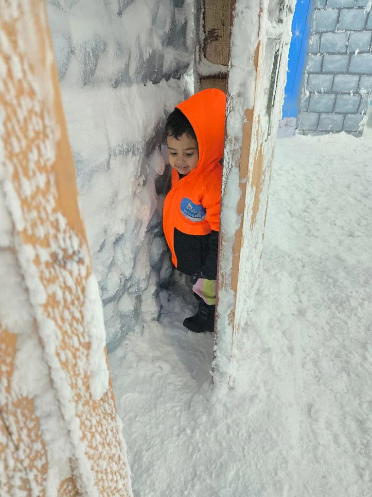 A young child peeks out from a snowy doorway, wearing a bright orange hooded jacket. The scene is a winter wonderland with snow-covered walls and ground.