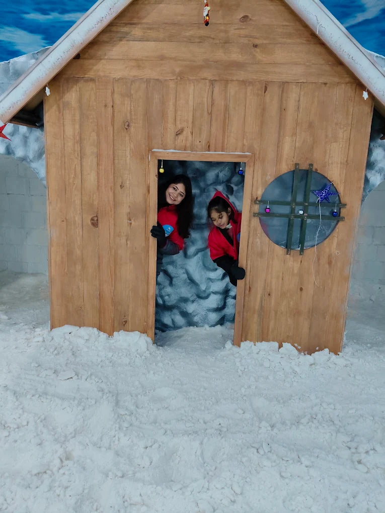 Two young girls in red outfits peek out of a wooden doorway in a snowy setting.