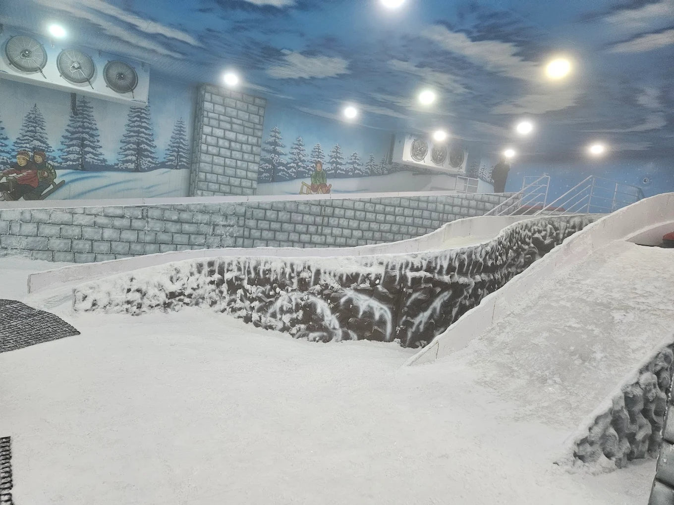 An indoor snow park with children sledding down snowy slopes under a painted sky.