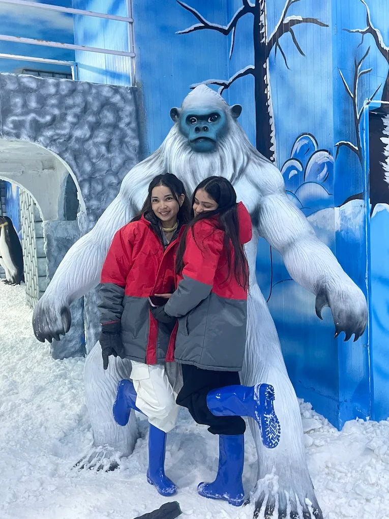 Two young women in winter jackets and boots pose with a large yeti statue in a snowy, blue-themed amusement park.