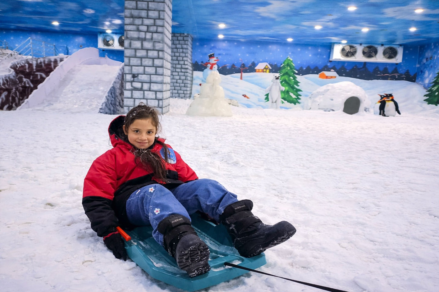 A young girl in a red jacket smiles while sitting on a sled in a snowy indoor park with cartoon penguins and a snowman.