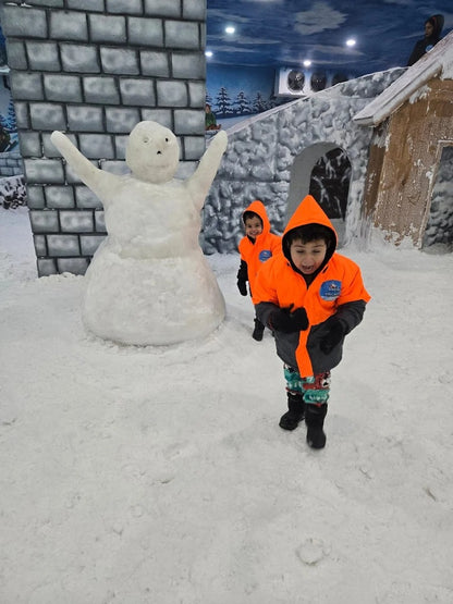 Two young children in orange jackets play in the snow next to a large snowman.
