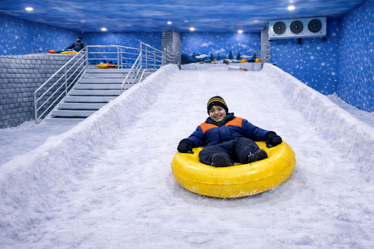 A young boy smiles while tubing down a snowy slope in an indoor winter wonderland.