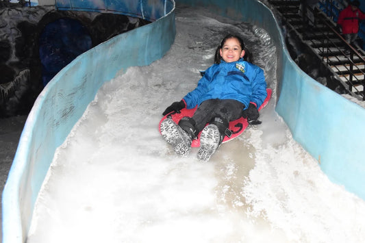 A young girl smiles as she slides down an icy slope on a red sled. She is wearing a blue jacket and black pants.