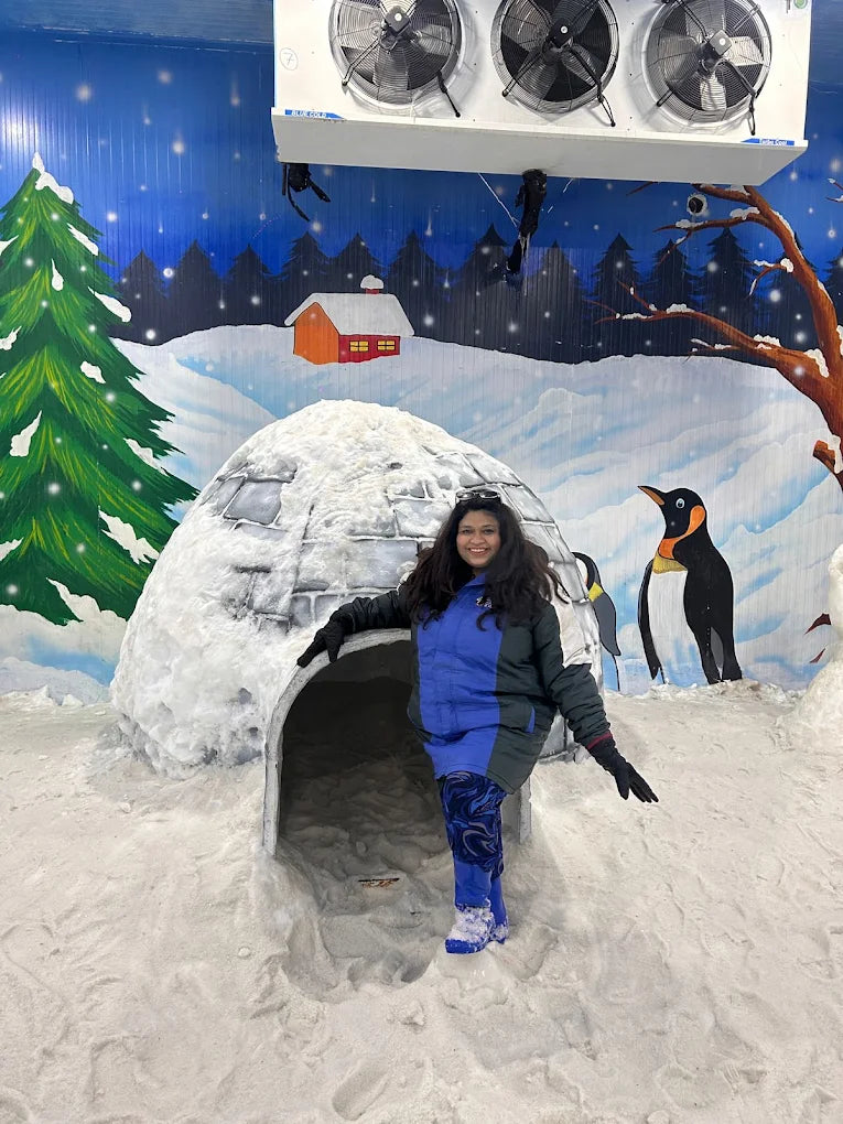 A woman stands in front of a snow igloo with painted penguins and trees in the background.