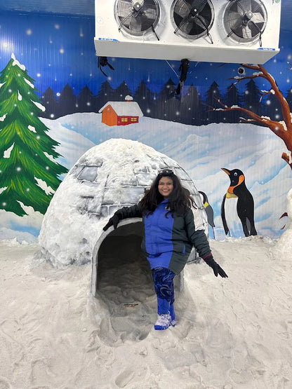 A woman stands in front of a snow igloo with painted penguins and trees in the background.
