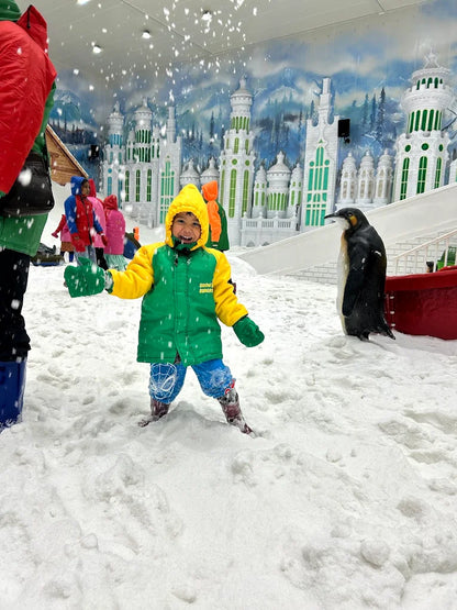 A young child in a snowsuit plays in the snow with a penguin statue and a snowy castle backdrop.